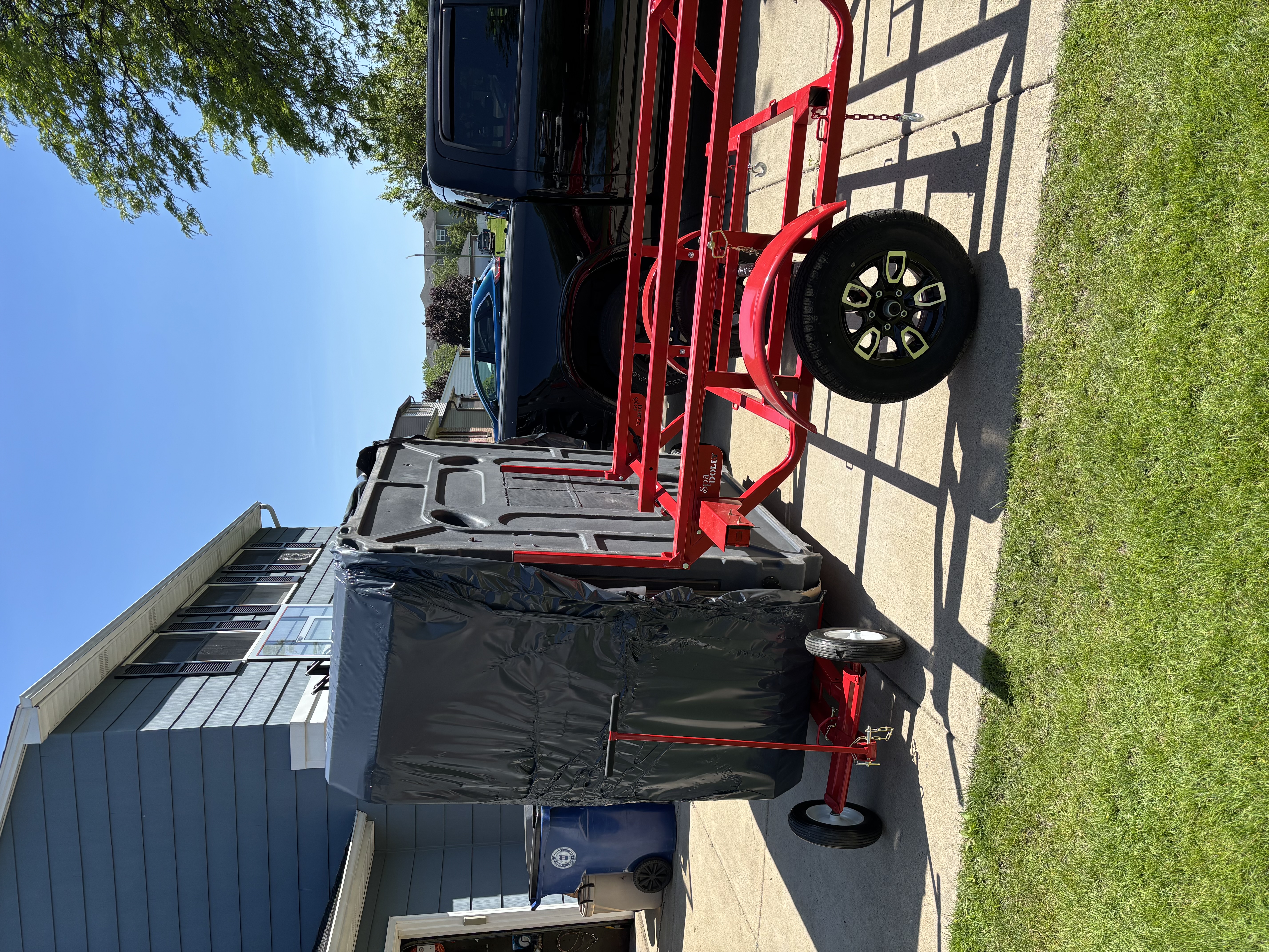 Wrapped hot tub standing upright on a red trailer in a residential driveway, ready for transport across Western New York